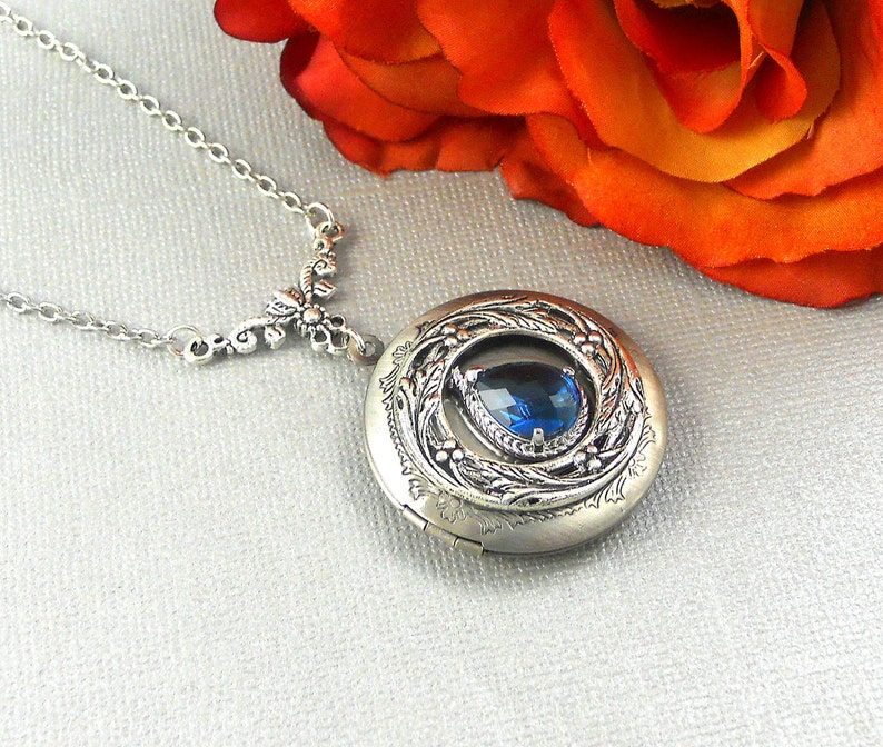 Puede incluir: Un collar de plata con un medall&oacute;n que tiene una piedra preciosa azul en forma de l&aacute;grima engastada en un dise&ntilde;o floral. El collar tiene una cadena delicada y un colgante floral.