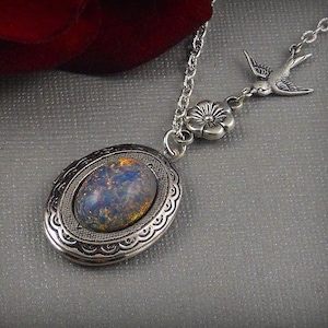 Puede incluir: Un collar de plata con un medallón que tiene una piedra de ópalo azul y dorada. El collar tiene una cadena fina de plata y un pequeño dije de flor de plata.