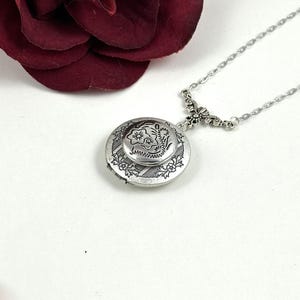 Può includere: Una collana d'argento con un ciondolo a forma di medaglione con un motivo floreale. Il medaglione è aperto e l'interno è visibile. La collana è su uno sfondo bianco.