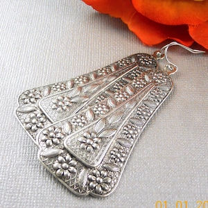 May include: Silver-coloured, fan-shaped earrings with intricate floral and leaf patterns. The earrings have a detailed, embossed design with a textured appearance. The hooks are visible at the top, and the earrings are displayed against a light background.
