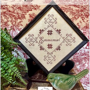 May include: A framed cross-stitch sampler featuring the word "Emmanuel" in the centre. The sampler is surrounded by four smaller squares with the words "King of Kings", "Lord of Lords", "Holy One", and "Mighty God".