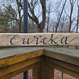 May include: A rustic wooden sign with the word "Eureka" in elegant black script. The sign is made from a piece of weathered wood, showcasing natural grain and texture. It rests on a wooden railing, with a blurred outdoor background.
