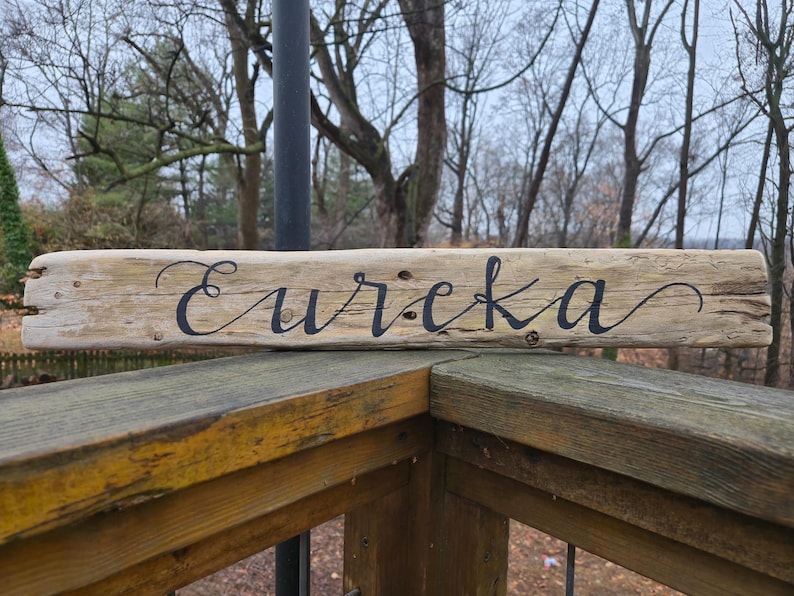 May include: A rustic wooden sign with the word "Eureka" in elegant black script. The sign is made from a piece of weathered wood, showcasing natural grain and texture. It rests on a wooden railing, with a blurred outdoor background.