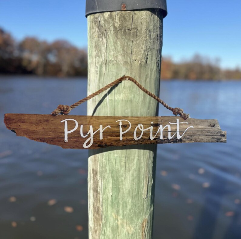 May include: A weathered wooden sign with the text "Pyr Point" in white script hangs from a rope. The sign is attached to a weathered wooden post. The background features a body of water and trees under a blue sky.