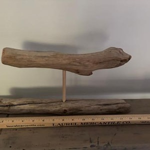 May include: A handcrafted driftwood sculpture. The sculpture features a horizontal piece of driftwood supported by a vertical wooden dowel, resting on a base of driftwood. A wooden ruler is in the foreground.
