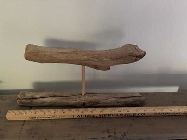 May include: A handcrafted driftwood sculpture. The sculpture features a horizontal piece of driftwood supported by a vertical wooden dowel, resting on a base of driftwood. A wooden ruler is in the foreground.
