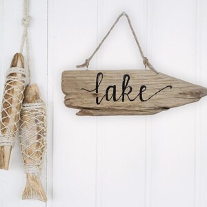 May include: Wooden sign with the word "lake" in black script, hanging from a natural rope. Two decorative wooden fish, wrapped in netting, hang from a rope on the left. The background is white.