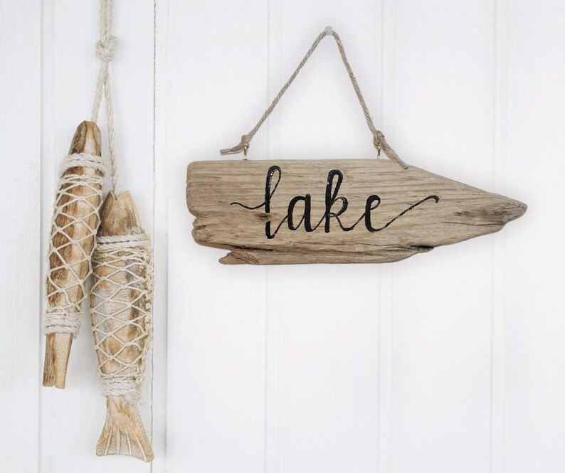 May include: Wooden sign with the word "lake" in black script, hanging from a natural rope. Two decorative wooden fish, wrapped in netting, hang from a rope on the left. The background is white.