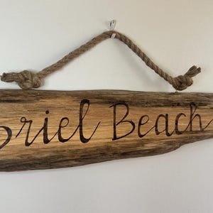May include: Wooden sign with the words "Briel Beach" in a cursive font. The sign is made of a piece of driftwood with a natural wood grain. A rope is attached for hanging.
