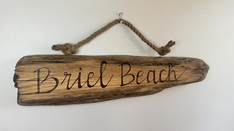 May include: Wooden sign with the words "Briel Beach" in a cursive font. The sign is made of a piece of driftwood with a natural wood grain. A rope is attached for hanging.