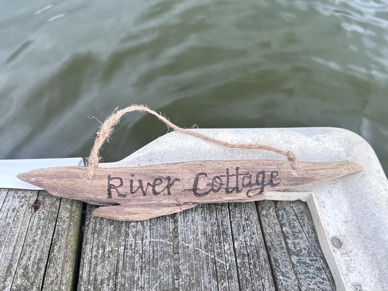 May include: A rustic wooden sign with the words "River Cottage" in black lettering. The sign is made from a piece of driftwood and is suspended by a natural twine. The background shows a body of water.
