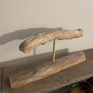 May include: A decorative driftwood sculpture. A long, light-colored piece of driftwood is supported by a wooden dowel, which is attached to a rectangular base of driftwood. The sculpture is displayed on a wooden shelf.