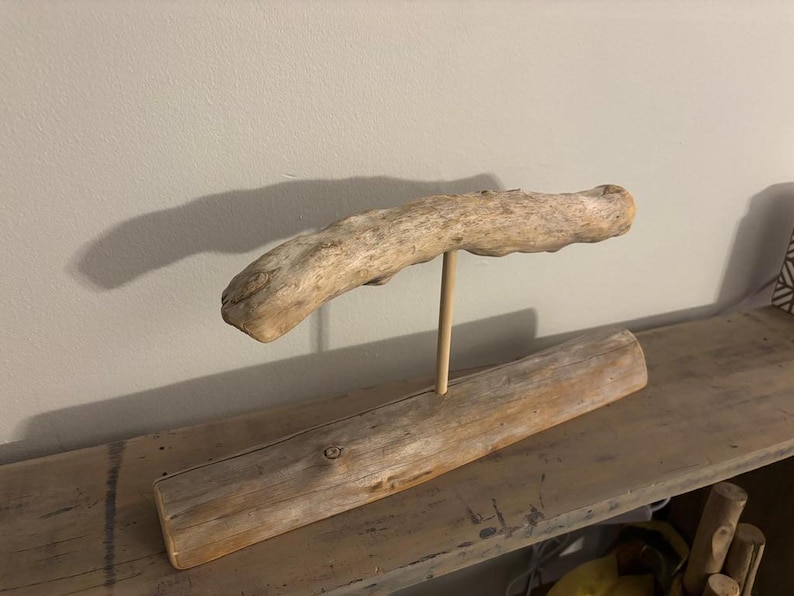 May include: A decorative driftwood sculpture. A long, light-colored piece of driftwood is supported by a wooden dowel, which is attached to a rectangular base of driftwood. The sculpture is displayed on a wooden shelf.