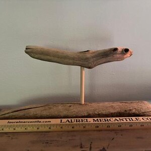 May include: A unique driftwood sculpture. The sculpture features a piece of driftwood supported by a wooden dowel, mounted on a larger piece of driftwood. A wooden ruler is in the foreground.