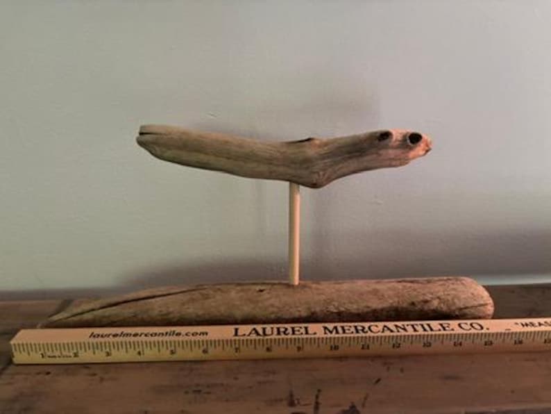 May include: A unique driftwood sculpture. The sculpture features a piece of driftwood supported by a wooden dowel, mounted on a larger piece of driftwood. A wooden ruler is in the foreground.