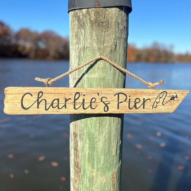May include: A wooden sign with the text "Charlie's Pier" in black script hangs from a rope. The sign is made of light-colored wood and is attached to a weathered green wooden post. The background shows a body of water and a blue sky.