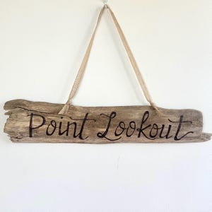 May include: A rustic wooden sign with the words "Point Lookout" in black script. The sign is made from a piece of driftwood and is suspended by a natural-colored ribbon. The sign is hanging against a white wall.