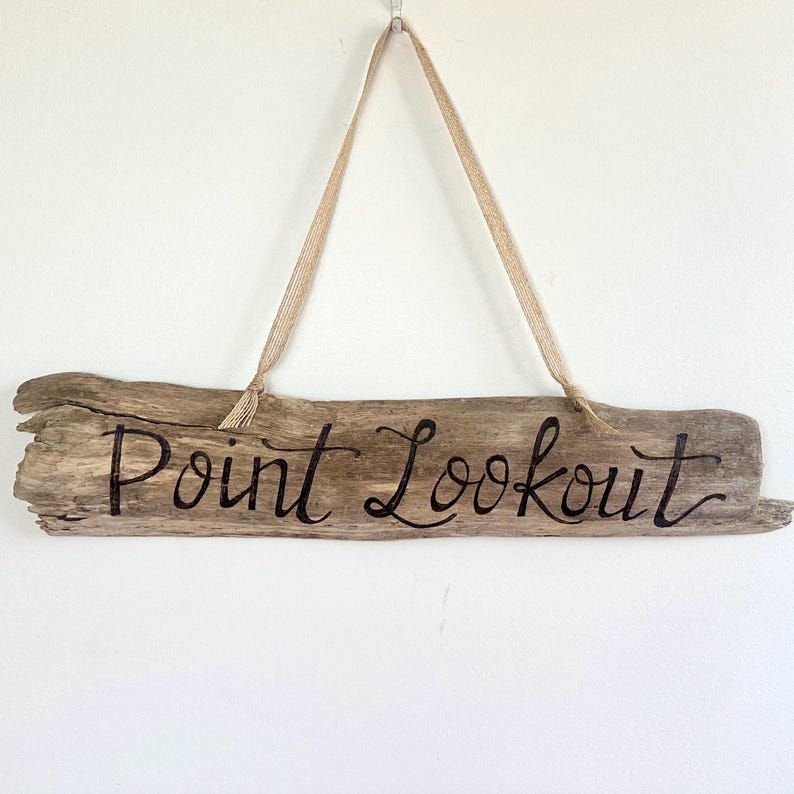 May include: A rustic wooden sign with the words "Point Lookout" in black script. The sign is made from a piece of driftwood and is suspended by a natural-colored ribbon. The sign is hanging against a white wall.