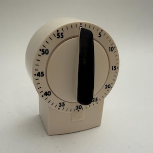 May include: A cream-colored kitchen timer with a black dial and a black handle. The timer has a circular face with black numbers indicating minutes, ranging from 5 to 55. The base is rectangular.