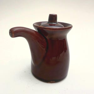 May include: A glossy, dark brown ceramic creamer with a curved spout and a small, round lid. The creamer has a bulbous base and a flared rim, and is likely vintage.