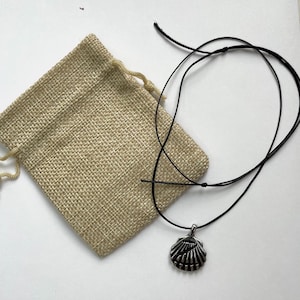 Puede incluir: Un collar con colgante de concha de color plateado con un cordón negro, junto a una bolsa de yute beige con cordón. El colgante es una réplica detallada de una concha. El collar se muestra sobre una superficie blanca.