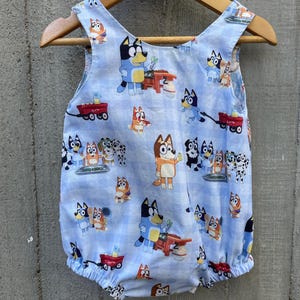 May include: A blue baby onesie with a pattern of cartoon dogs. The dogs are wearing different colored shirts and pants. The onesie has a red wagon and other details on it.