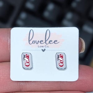 May include: A pair of stud earrings shaped like miniature silver Diet Coke cans with red and white branding. The earrings are attached to a white rectangular display card featuring the text lovelee Low Co. with a small heart icon centered below.
