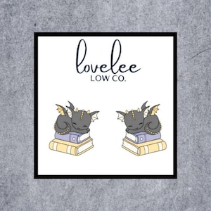 May include: Square graphic with the text "lovelee LOW CO." above two illustrations of sleeping gray dragons atop stacks of books. The dragons have yellow wings and horns. The books are yellow and purple.
