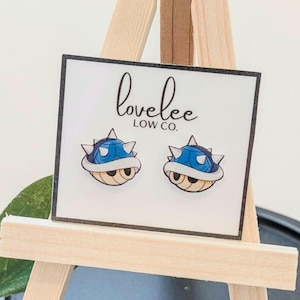May include: A pair of blue and white cartoon turtle shell earrings on a white card. The card has the text "lovelee LOW CO." in black script. The earrings are displayed on a small wooden easel.