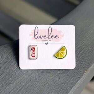 May include: A pair of stud earrings on a white display card labeled lovelee Low Co. One earring is a miniature silver and red Diet Coke soda can, and the other earring is a yellow and green lime wedge. The earrings are small and detailed.