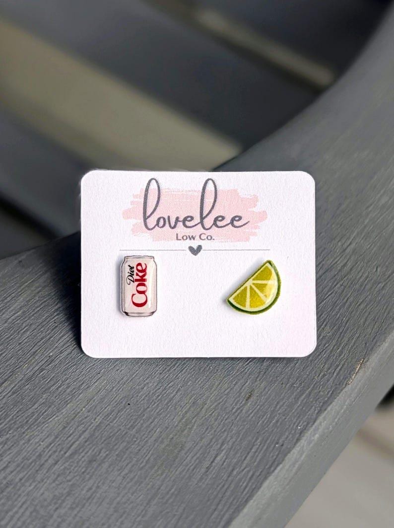 May include: A pair of stud earrings on a white display card labeled lovelee Low Co. One earring is a miniature silver and red Diet Coke soda can, and the other earring is a yellow and green lime wedge. The earrings are small and detailed.