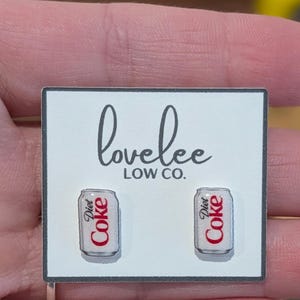 May include: A pair of stud earrings shaped like silver soda cans with the red and black Diet Coke logo printed on the front. The earrings are attached to a white rectangular display card featuring the black text lovelee LOW CO. at the top.