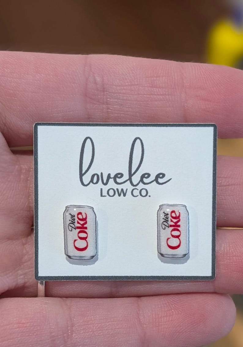 May include: A pair of stud earrings shaped like silver soda cans with the red and black Diet Coke logo printed on the front. The earrings are attached to a white rectangular display card featuring the black text lovelee LOW CO. at the top.