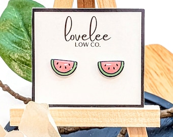 Könnte beinhalten: Ohrringe mit Wassermelonen-Scheiben auf einer weißen Karte mit dem Text "lovelee LOW CO.". Die Ohrringe sind rosa mit schwarzen Samen und einer grünen Schale. Die Karte wird auf einer kleinen Holzstaffelei präsentiert.