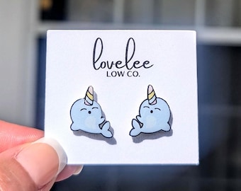 May include: A pair of blue narwhal stud earrings with yellow and pink horns. The earrings are on a white card with the text "lovelee LOW CO."