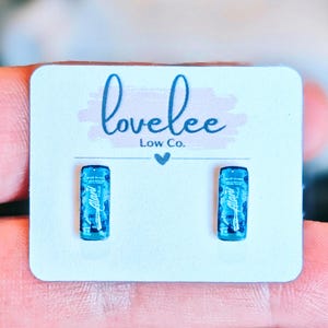 May include: A pair of small stud earrings shaped like miniature blue energy drink cans. The earrings are attached to a white display card featuring the text "lovelee Low Co." in blue script with a small heart icon centered below the brand name.