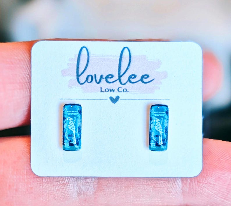 May include: A pair of small stud earrings shaped like miniature blue energy drink cans. The earrings are attached to a white display card featuring the text "lovelee Low Co." in blue script with a small heart icon centered below the brand name.