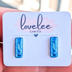 May include: A pair of stud earrings featuring miniature blue energy drink cans with the text Alani Energy printed on the front. The earrings are attached to a white rectangular display card with the text lovelee Low Co. printed in black.