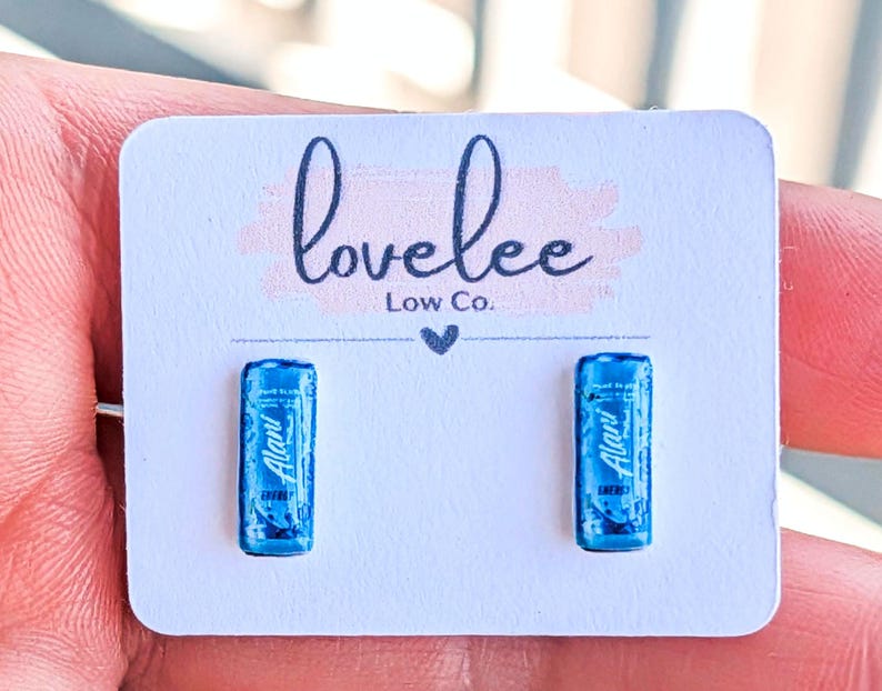 May include: A pair of stud earrings featuring miniature blue energy drink cans with the text Alani Energy printed on the front. The earrings are attached to a white rectangular display card with the text lovelee Low Co. printed in black.