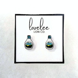 May include: A pair of novelty earrings featuring miniature ranch dressing bottles. The earrings are white with blue and green accents, and the word "RANCH" is visible on the label. The earrings are displayed on a white card with the text "lovelee LOW CO."
