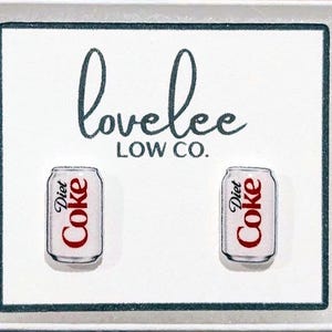 May include: A pair of Diet Coke can earrings. The earrings are white with red text that reads "Diet Coke". The earrings are on a white background with the text "lovelee LOW CO." in black script.