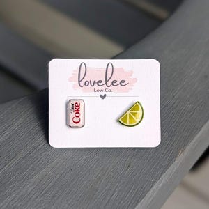 May include: A pair of stud earrings on a white display card labeled lovelee Low Co. One earring is a miniature silver and red Diet Coke soda can, and the other is a yellow and green lime wedge slice. The earrings are small and detailed.