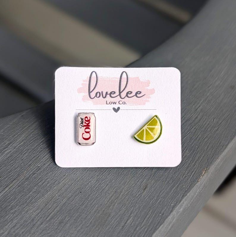 May include: A pair of stud earrings on a white display card labeled lovelee Low Co. One earring is a miniature silver and red Diet Coke soda can, and the other is a yellow and green lime wedge slice. The earrings are small and detailed.