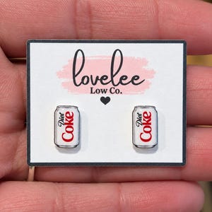 May include: A pair of stud earrings shaped like miniature silver and white Diet Coke soda cans with red lettering. The earrings are displayed on a white rectangular card featuring the black text lovelee Low Co. and a small black heart icon.