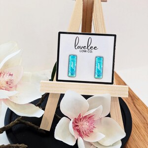May include: A pair of rectangular stud earrings featuring a bright turquoise graphic design with the text Alani printed on each. The earrings are displayed on a white card labeled lovelee LOW CO. and are mounted on a small wooden easel stand.