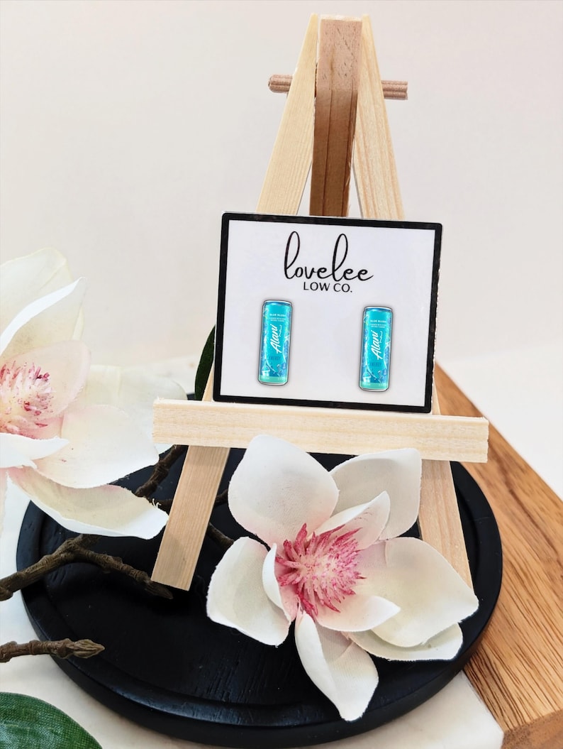 May include: A pair of rectangular stud earrings featuring a bright turquoise graphic design with the text Alani printed on each. The earrings are displayed on a white card labeled lovelee LOW CO. and are mounted on a small wooden easel stand.