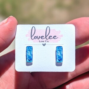 May include: A pair of small, rectangular stud earrings featuring a miniature blue energy drink can design with the text Alani Nu printed on the front. The earrings are attached to a white display card with the text lovelee Low Co. printed in black.