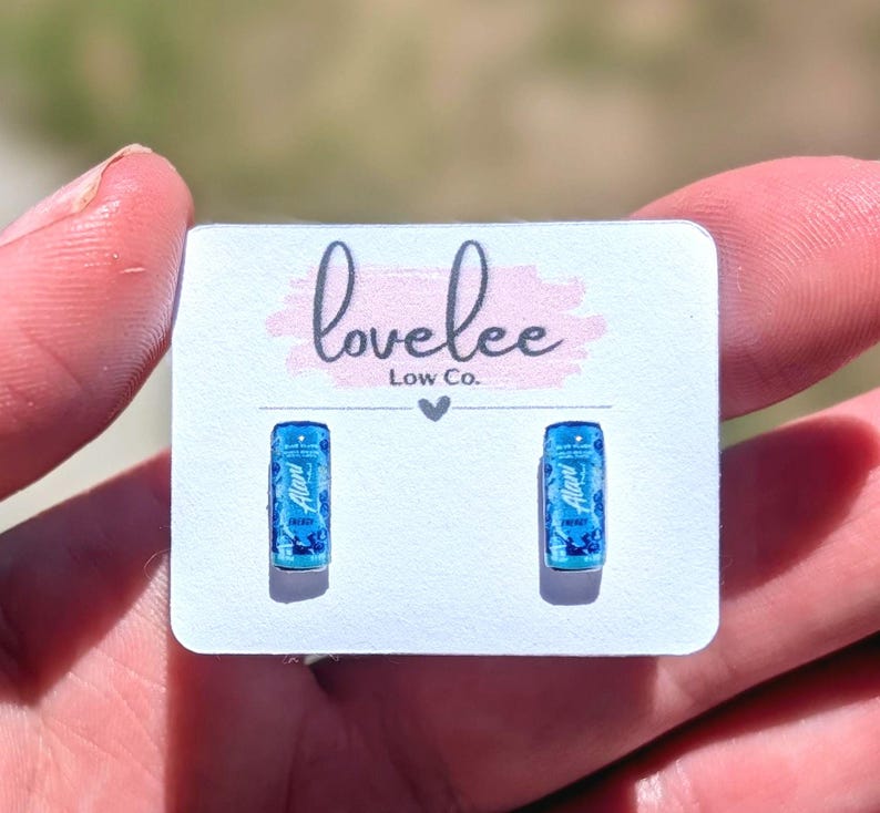 May include: A pair of small, rectangular stud earrings featuring a miniature blue energy drink can design with the text Alani Nu printed on the front. The earrings are attached to a white display card with the text lovelee Low Co. printed in black.