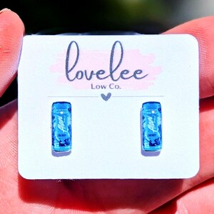 May include: A pair of stud earrings shaped like miniature blue energy drink cans, each featuring the Alani Nu logo and branding. The earrings are mounted on a white display card printed with the text lovelee Low Co. and a small heart icon.