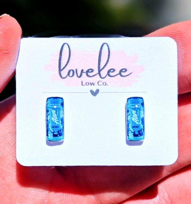 May include: A pair of stud earrings shaped like miniature blue energy drink cans, each featuring the Alani Nu logo and branding. The earrings are mounted on a white display card printed with the text lovelee Low Co. and a small heart icon.